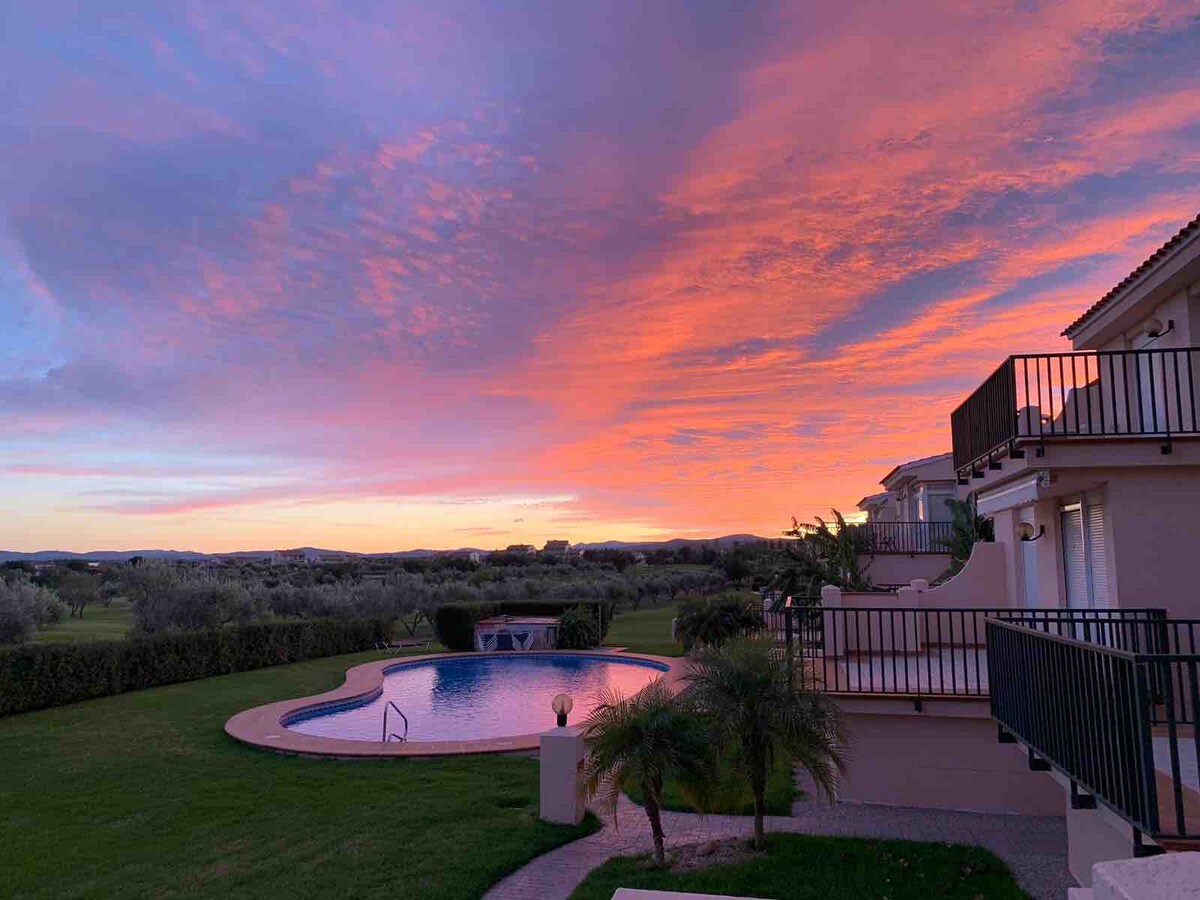 A serene view showcases the swimming pool surrounded by lush grass, reflecting the vibrant colors of a sunset sky. The backdrop features the rolling landscape of a golf course and olive trees, enhancing the peaceful atmosphere.