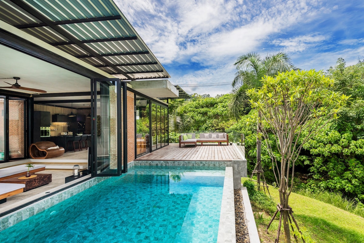 An infinity pool glistens under a clear blue sky, surrounded by lush greenery. Large glass doors are visible, leading to an open living area. A wooden deck features lounge seating, blending seamlessly with the natural landscape.