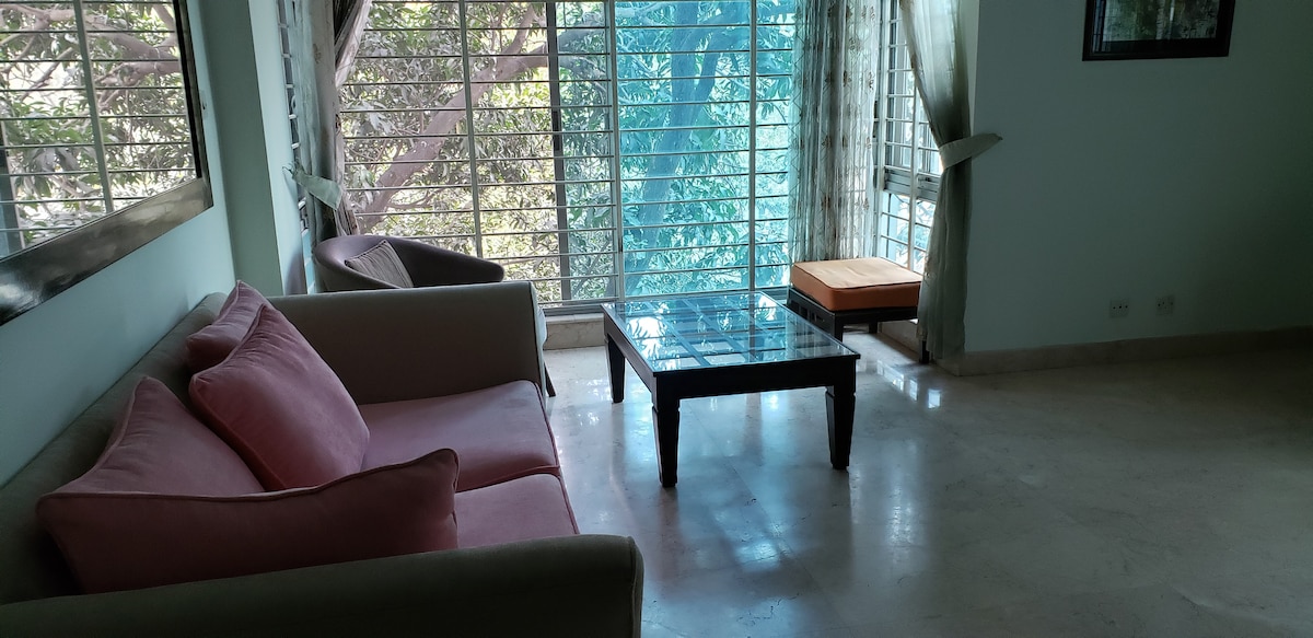 A comfortable living area is presented with a light-colored couch featuring pink cushions. A glass coffee table rests on the elegantly tiled floor, while sunlight filters through partially opened window panes, illuminating the space. Two armchairs are positioned to create a cozy seating arrangement.