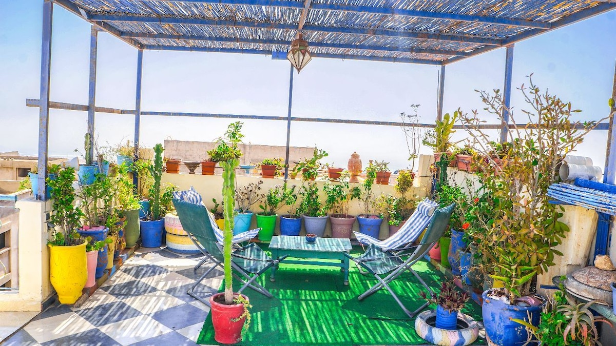 A spacious rooftop terrace is adorned with various potted plants, creating a vibrant green space. Two lounge chairs are positioned on a green carpet, inviting relaxation. A shaded area above provides comfort, while the distant view offers a glimpse of the surrounding environment.