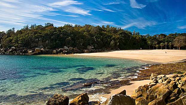 A sandy beach is framed by lush greenery, with calm turquoise waters gently lapping against the shore. Rocky formations create a natural boundary along the water's edge, while a clear blue sky stretches overhead, adorned with wispy clouds.