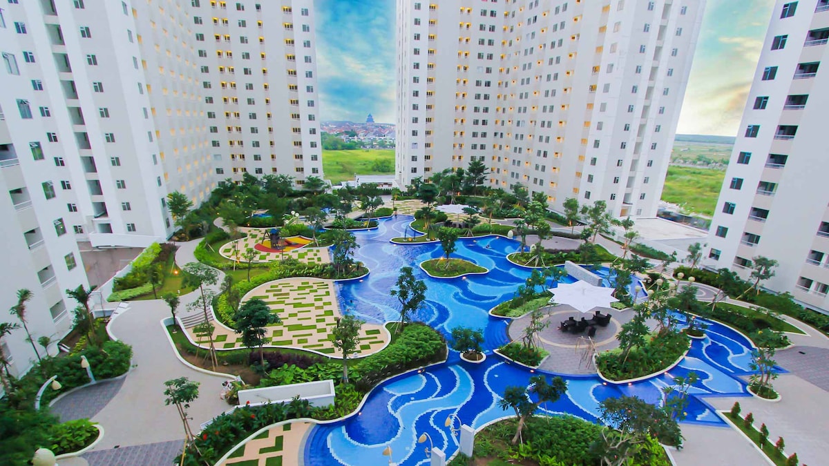 An aerial view showcases the lush landscape surrounding a swimming pool area, featuring meandering water pathways. Towering white buildings frame the vibrant outdoor space, which is decorated with greenery and seating areas for relaxation.