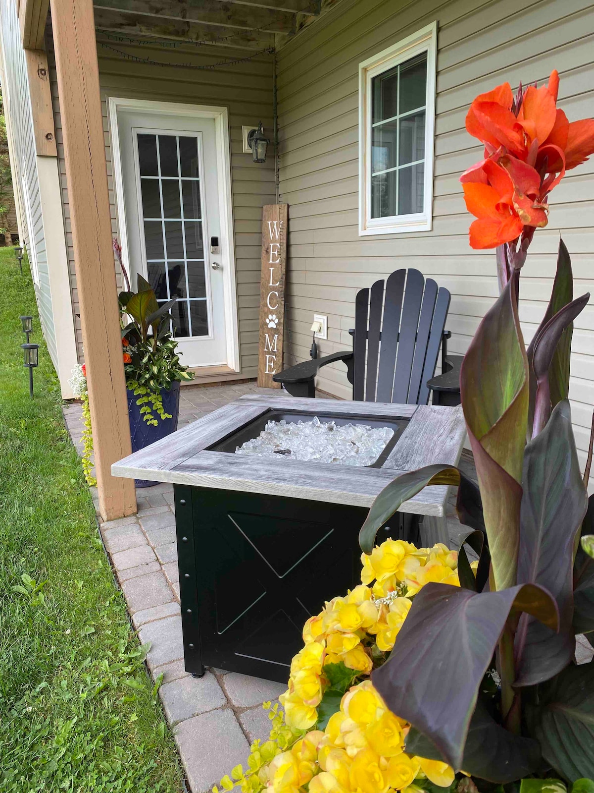 An outdoor seating area is visible with an Adirondack chair positioned near a fire table topped with glass stones. Colorful flowers, including yellow orchids and tall, vibrant stalks, frame the space. A welcoming sign and a door lead to the suite's entrance, indicating a homey atmosphere.