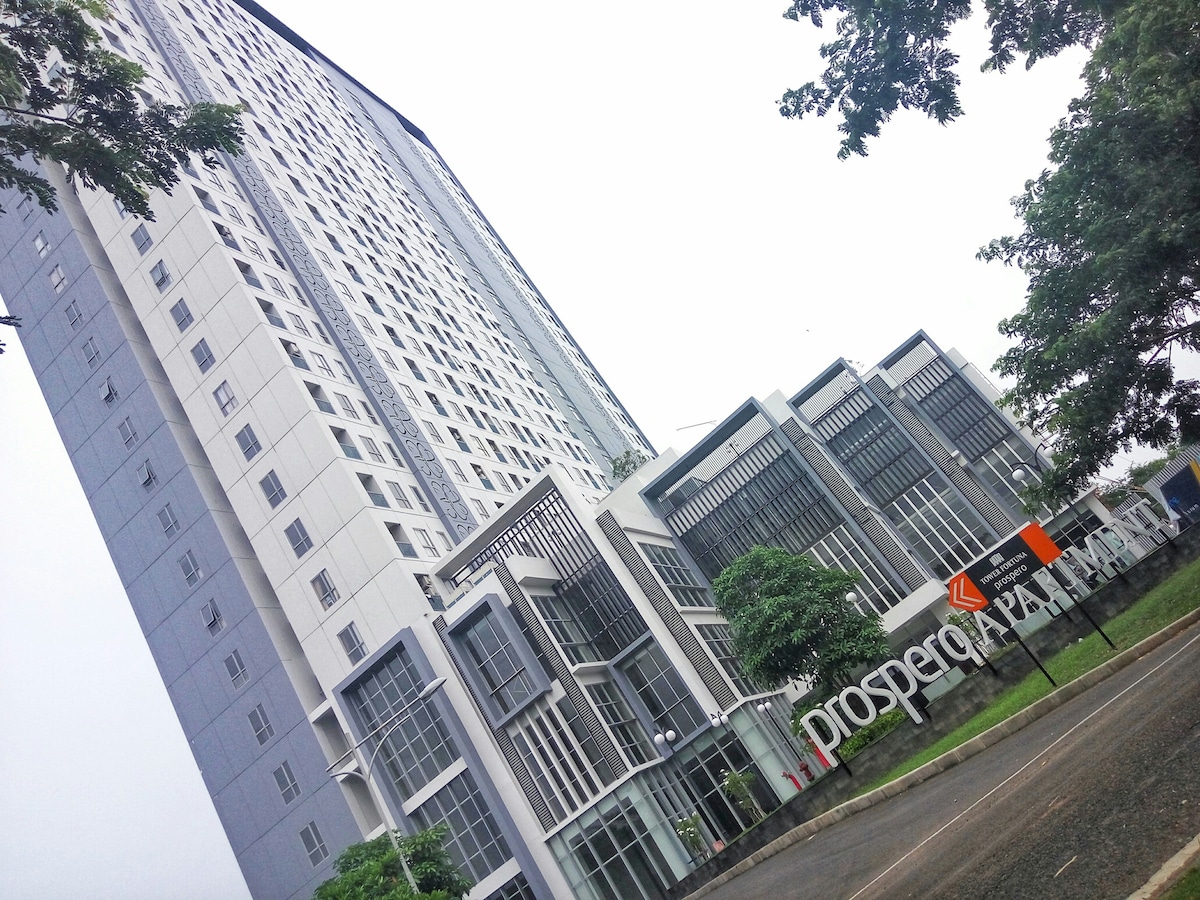 A modern apartment building is showcased, featuring a sleek facade with large windows and balconies. The entrance is marked by a prominent sign that reads 'Prospera' and is surrounded by landscaped greenery, creating a welcoming first impression.