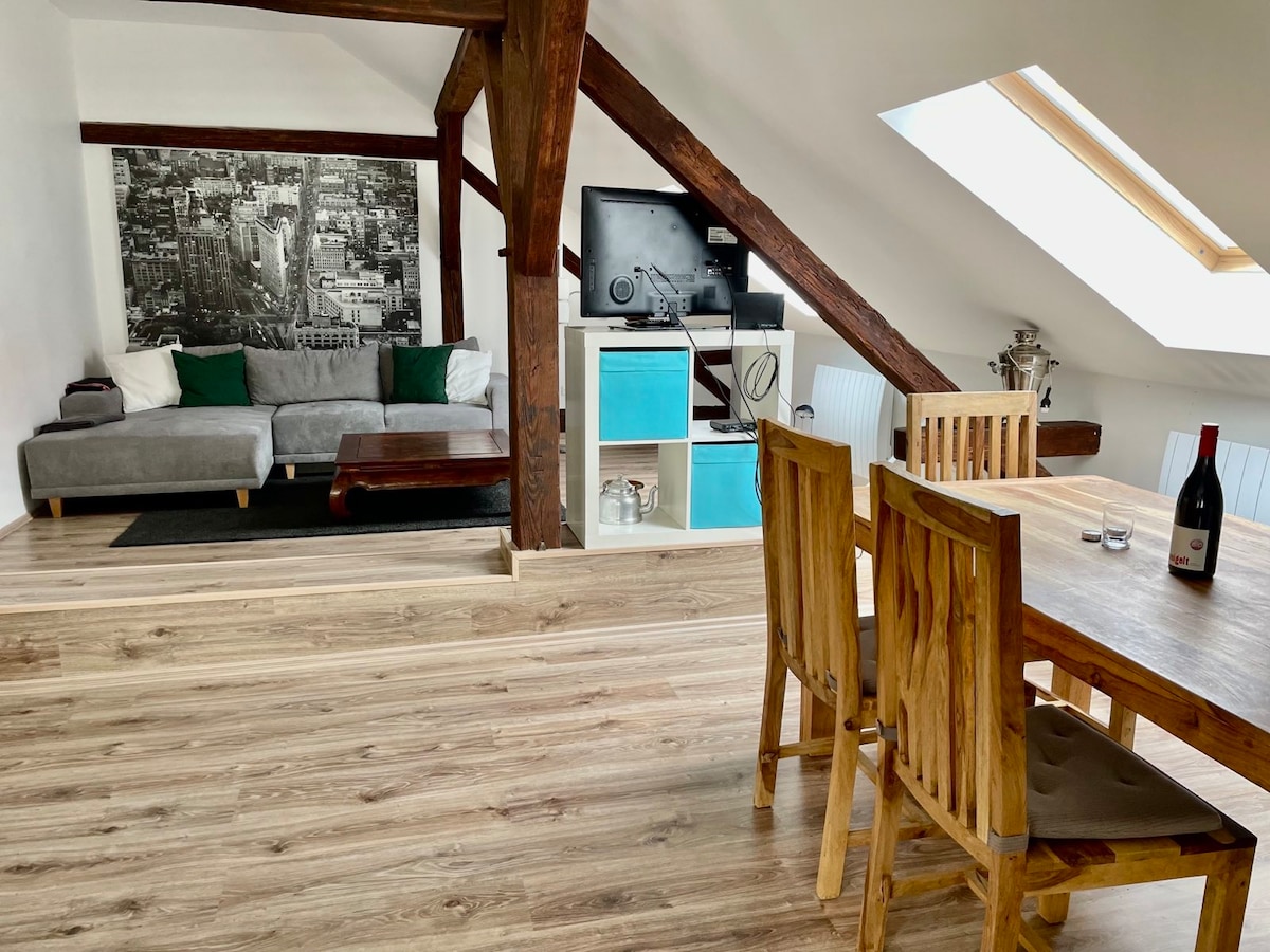 A spacious living area is presented with a comfortable gray sofa adorned with green cushions, situated next to a coffee table. A wooden dining table with four chairs occupies one corner, while a large artwork and a TV serve as focal points along the wall. Natural light filters through a skylight.