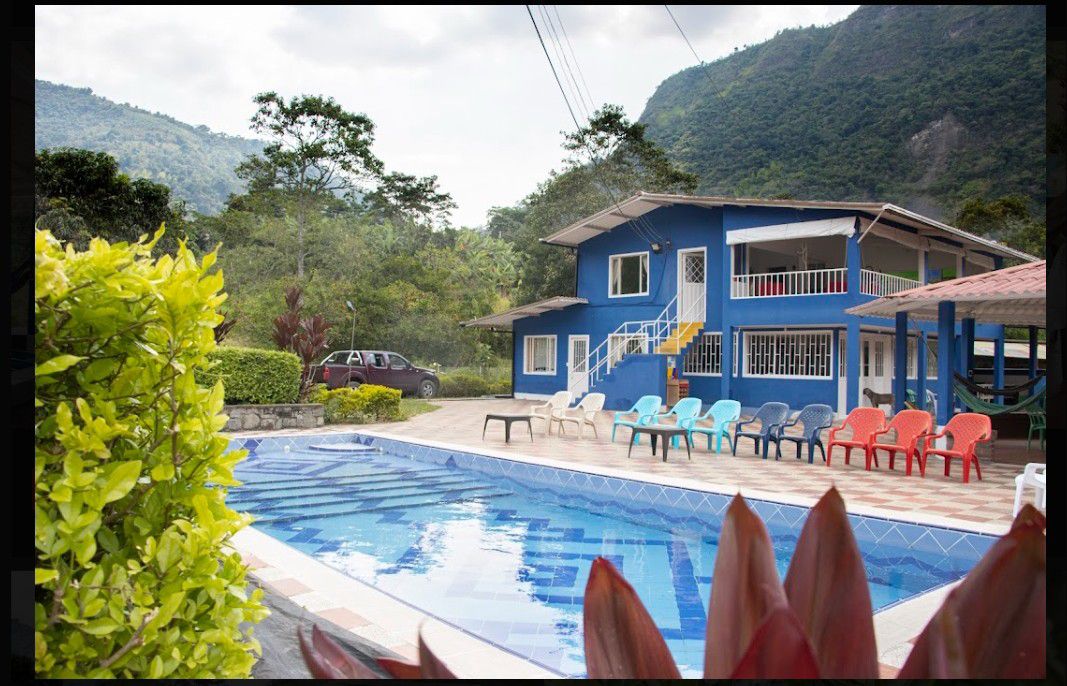 A vibrant blue house with a spacious deck overlooks a well-maintained pool. Lounge chairs in various colors are arranged around the pool area, with lush greenery and mountains providing a serene backdrop.