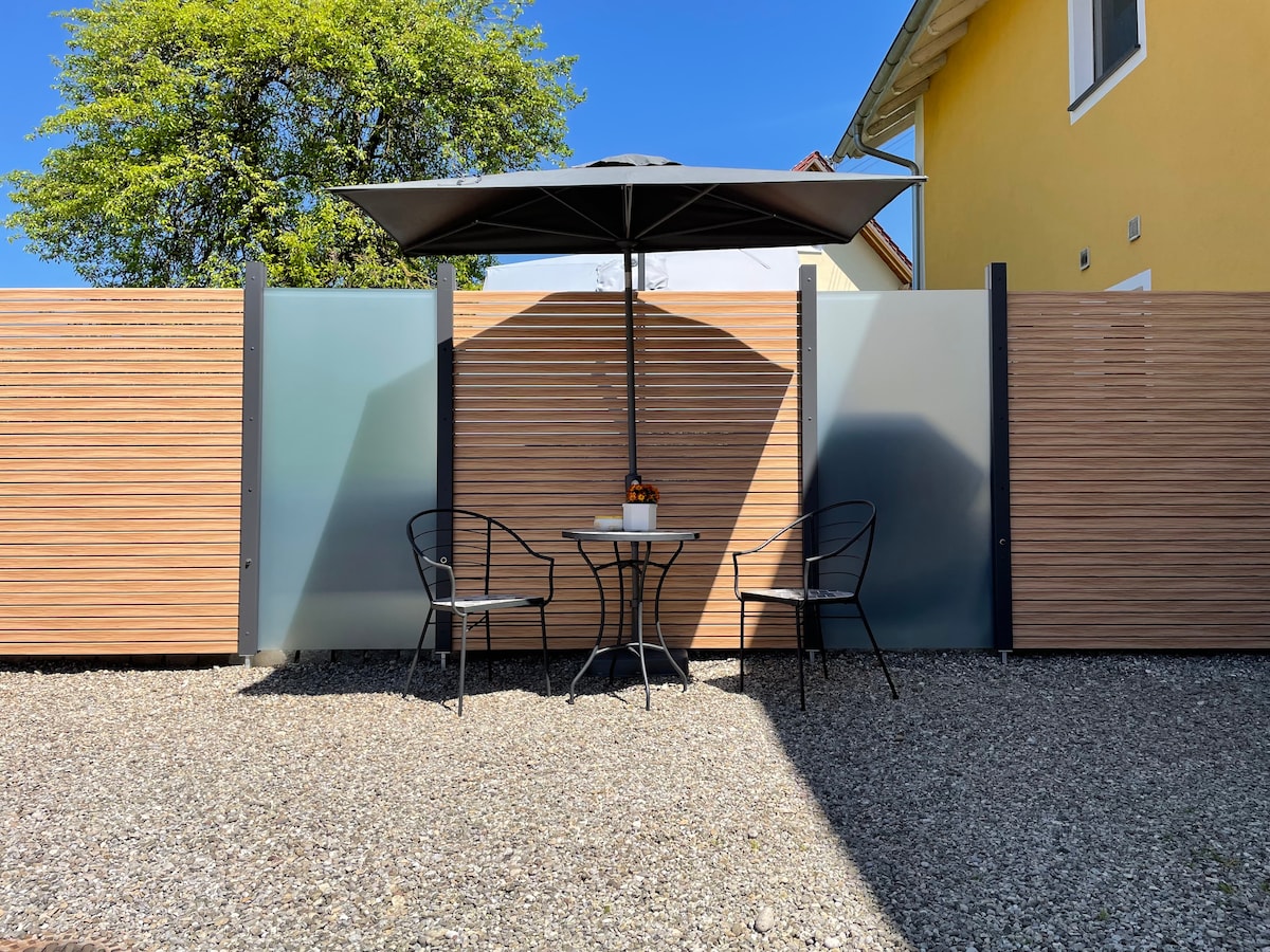 A shaded outdoor seating area features a round table with a small decorative centerpiece. Two black metal chairs are positioned around the table. A large umbrella provides shade, while low wooden fencing and a gravel surface create a relaxed, private ambiance.