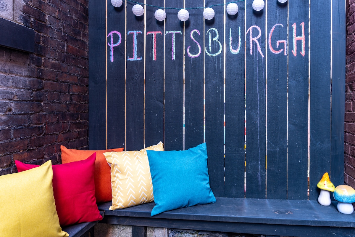 A vibrant outdoor seating area features a rustic wooden wall painted with the word 'Pittsburgh.' Colorful cushions in orange, yellow, and turquoise add comfort to the sleek bench below. Decorative string lights hang above, illuminating the space with a warm glow.