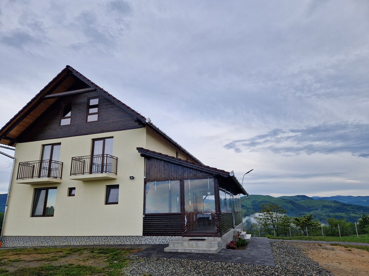A two-story cabin is presented, showcasing a combination of light-colored walls and dark wooden accents. Balconies are visible on the upper floor, while a spacious glass-enclosed area extends from the ground floor. Surrounding mountains contribute to a serene backdrop.