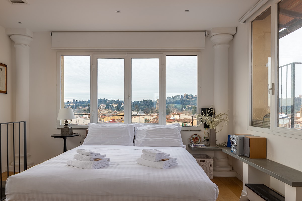 A spacious bedroom features a large bed dressed in crisp white linens. Tall windows offer a panoramic view of Florence, showcasing rooftops and greenery. A bedside table and a minimalist desk are visible, contributing to a clean and modern aesthetic.