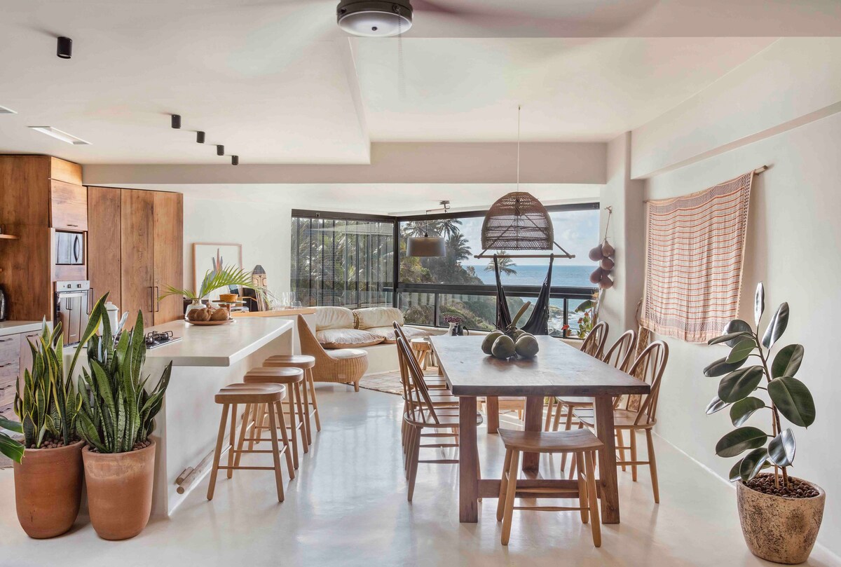 A bright and airy kitchen and dining area is showcased, with a large wooden dining table surrounded by light-colored chairs. Natural plants are present in decorative pots, and large windows provide a view of the ocean, enhancing the space with natural light.