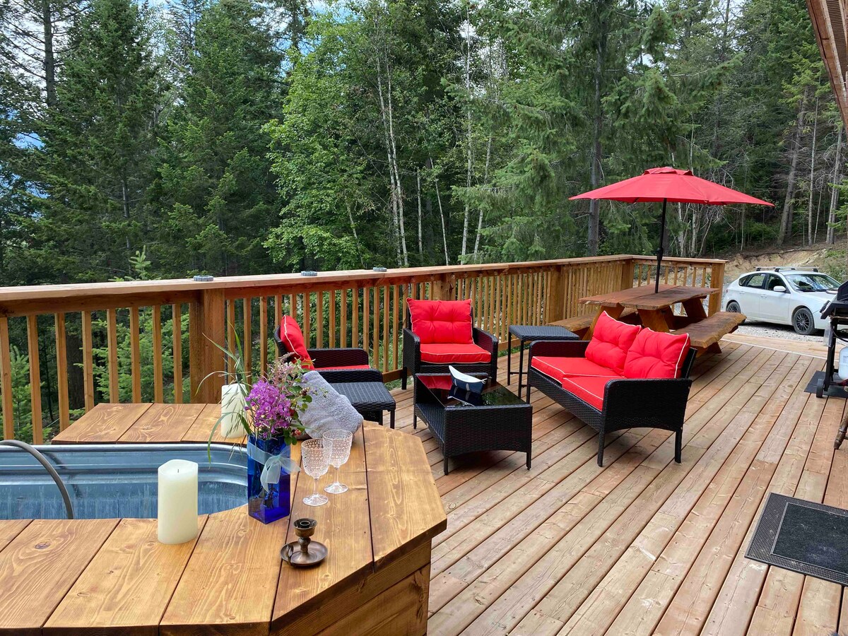 A spacious deck is highlighted by a rustic wooden hot tub, surrounded by comfortable seating with vibrant red cushions. A picnic table is positioned nearby, and an umbrella provides shade. Tall trees create a natural backdrop, enhancing the serene outdoor setting.