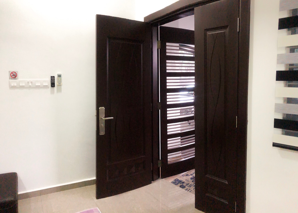 The entrance area displays a set of double doors with a dark wood finish. The doors are opened to reveal an interior space, featuring a tiled floor and a decorative mat. Adjacent walls are painted white, enhancing the bright and welcoming atmosphere.