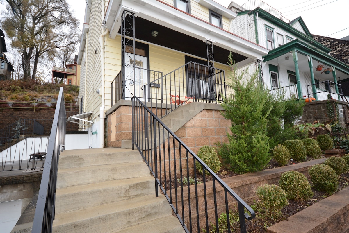 The exterior of a two-story house is visible, featuring a welcoming front porch with a railing. Stairs lead up from the sidewalk to the entrance. Neatly trimmed bushes line the walkway, and residences in varying colors are seen beside the house.