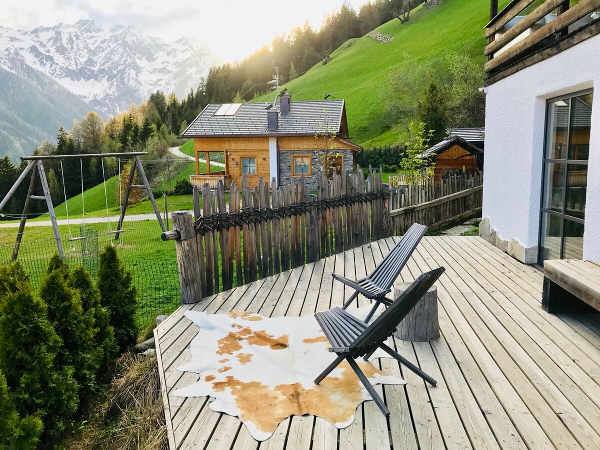 A wooden deck features two black lounge chairs positioned beside a decorative cowhide rug. A rustic fence borders the deck, while a grassy hillside and wooden structures are visible in the background, framed by snow-capped mountains under a soft sunset.