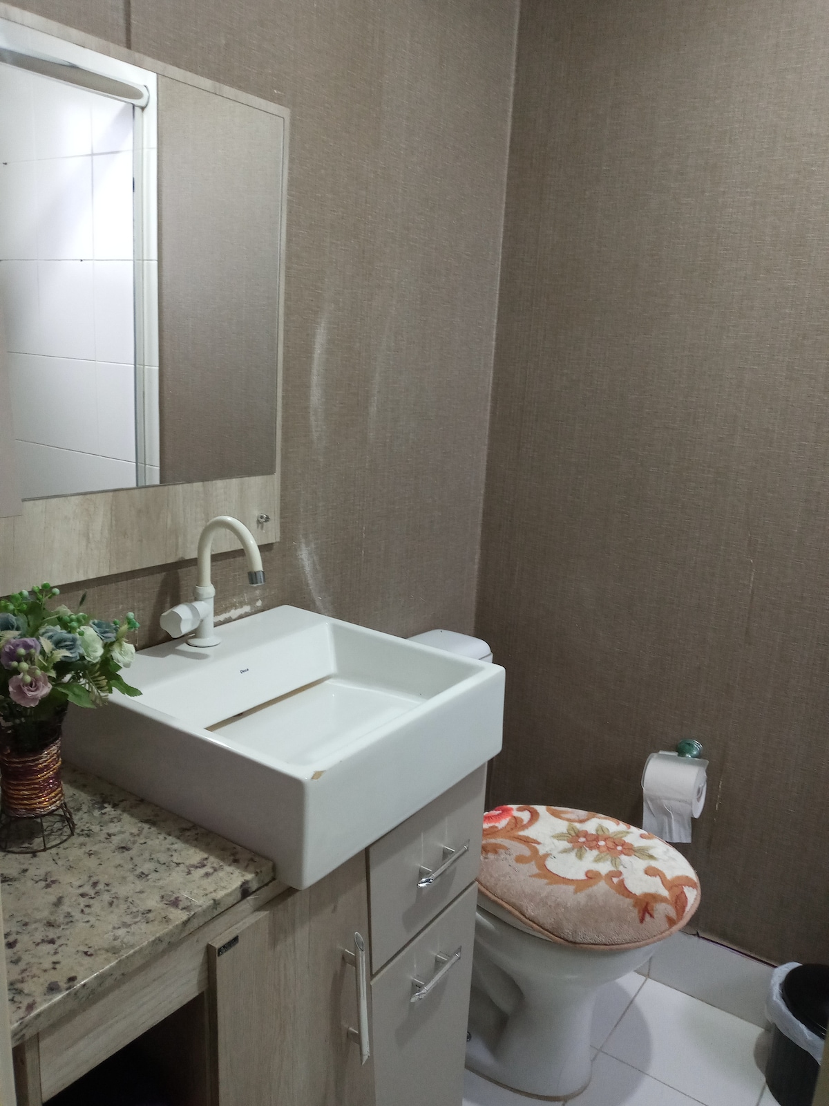 The bathroom features a minimalist design with textured wall finishes. A sleek countertop with an integrated sink is positioned alongside a modern mirror. The toilet is complemented by a decorative seat cover, while a small vase with flowers adds a touch of color.