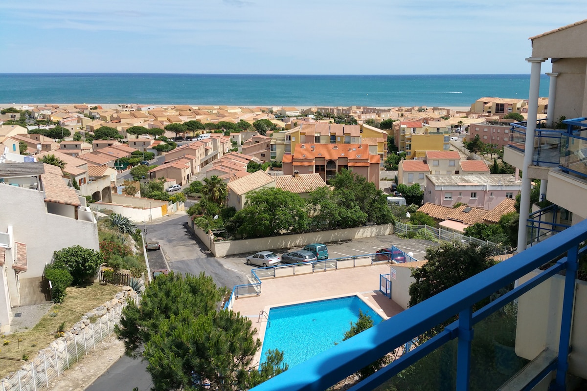 A panoramic view reveals the tranquil sea, with residential buildings and rooftops visible below. The scene includes a sparkling blue swimming pool surrounded by greenery, offering a serene atmosphere. Light clouds drift across a bright sky, enhancing the peaceful coastal setting.