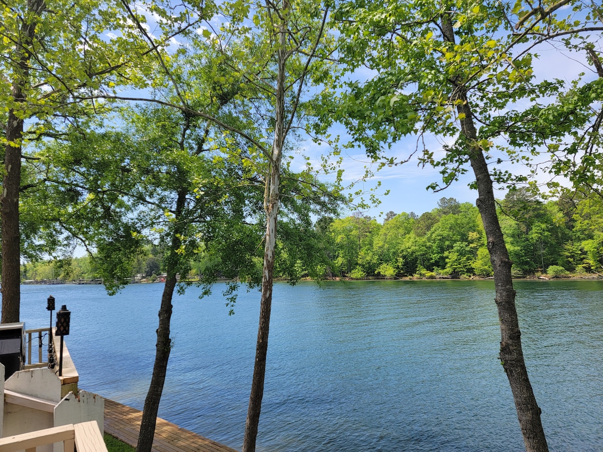 Tall trees frame a serene view of the lake, with sunlight reflecting on the calm water. Lush greenery borders the shoreline, creating a peaceful natural setting that invites relaxation and enjoyment of the outdoors.