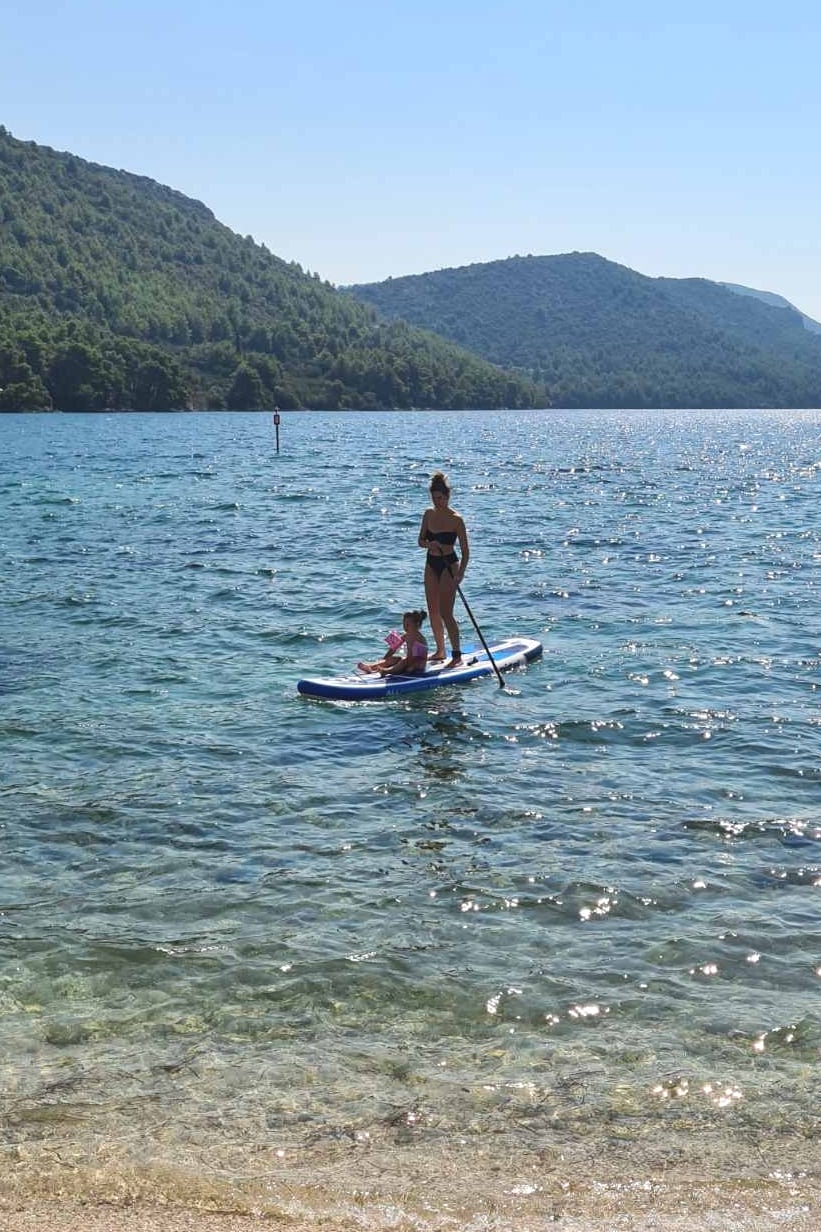 A paddleboard is viewed gliding over calm waters, with two individuals standing on it. The serene landscape features lush green hills rising from the water's edge, offering a scenic backdrop beneath a clear blue sky.