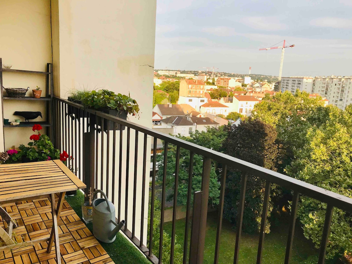 A balcony is presented with a wooden table and two chairs, overlooking a lush green area. Flowering plants are arranged along the railing, while a watering can sits on the floor. In the background, residential buildings and a construction crane can be seen against a clear sky.