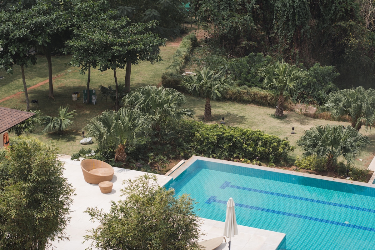 An expansive swimming pool is set against a backdrop of lush greenery, featuring palm trees and well-maintained lawns. The pool area includes a sunshade and round seating, inviting relaxation. The serene view encompasses a blend of natural landscaping and tranquil water.