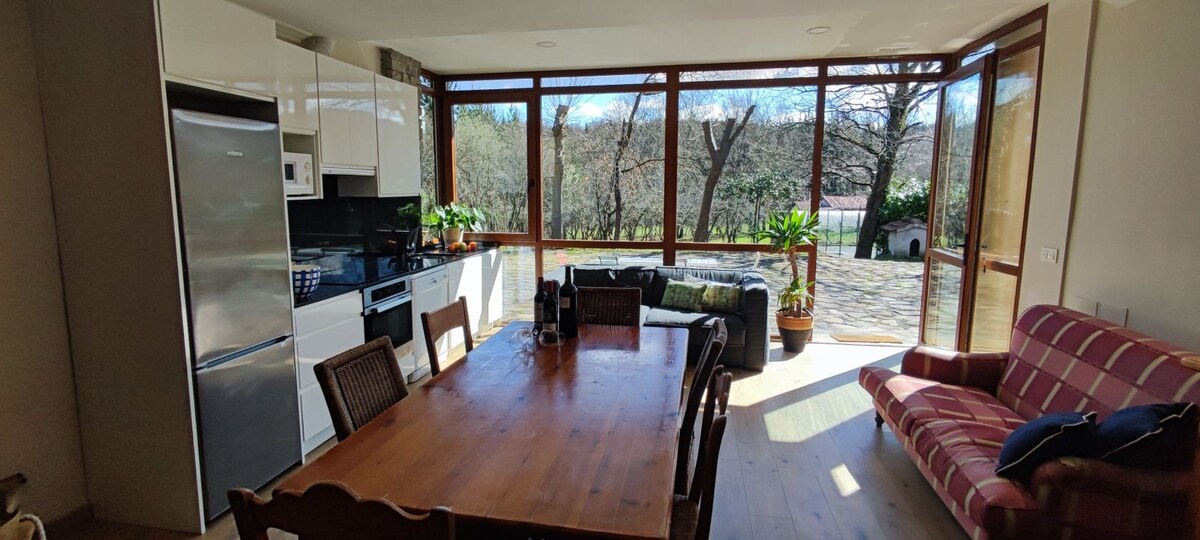 A spacious living area is presented with large windows that provide natural light and views of the surrounding nature. An expansive wooden dining table sits in the center, accompanied by a mix of chairs. The kitchen area, featuring modern appliances, is integrated into the open layout.