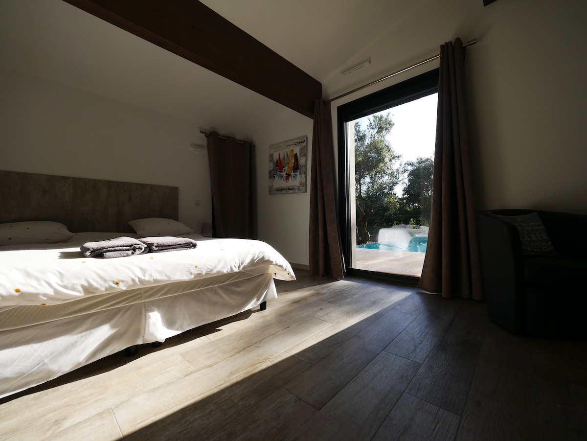 A spacious bedroom features a large bed with light linens and dark towels neatly arranged. Floor-to-ceiling glass doors open to a view of the pool and exterior greenery. Natural light floods the room, while dark curtains frame the doorway.
