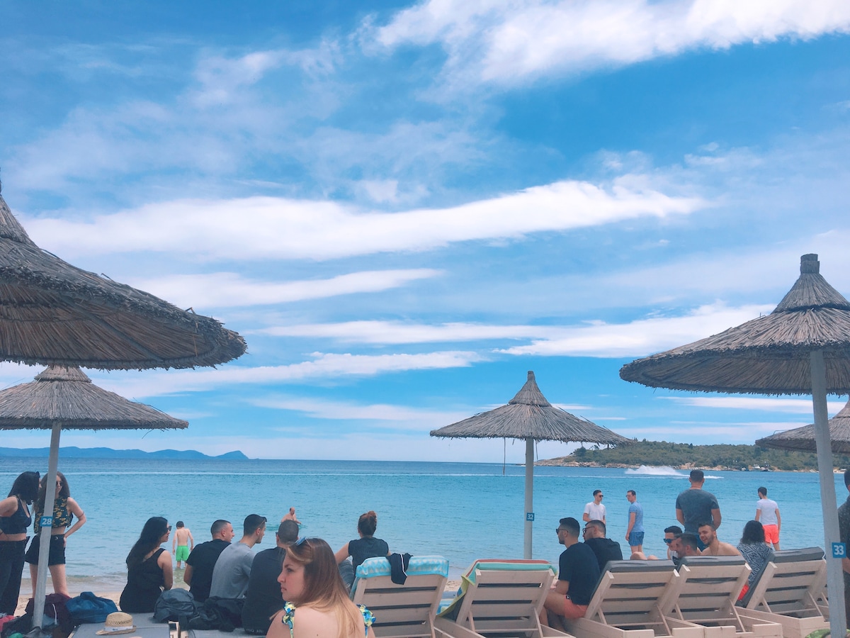 A lively beach scene features several sun loungers under thatched umbrellas, with groups of people relaxing and enjoying the shoreline. Calm blue waters stretch towards the horizon, while fluffy clouds drift across a bright sky, creating a relaxed coastal atmosphere.