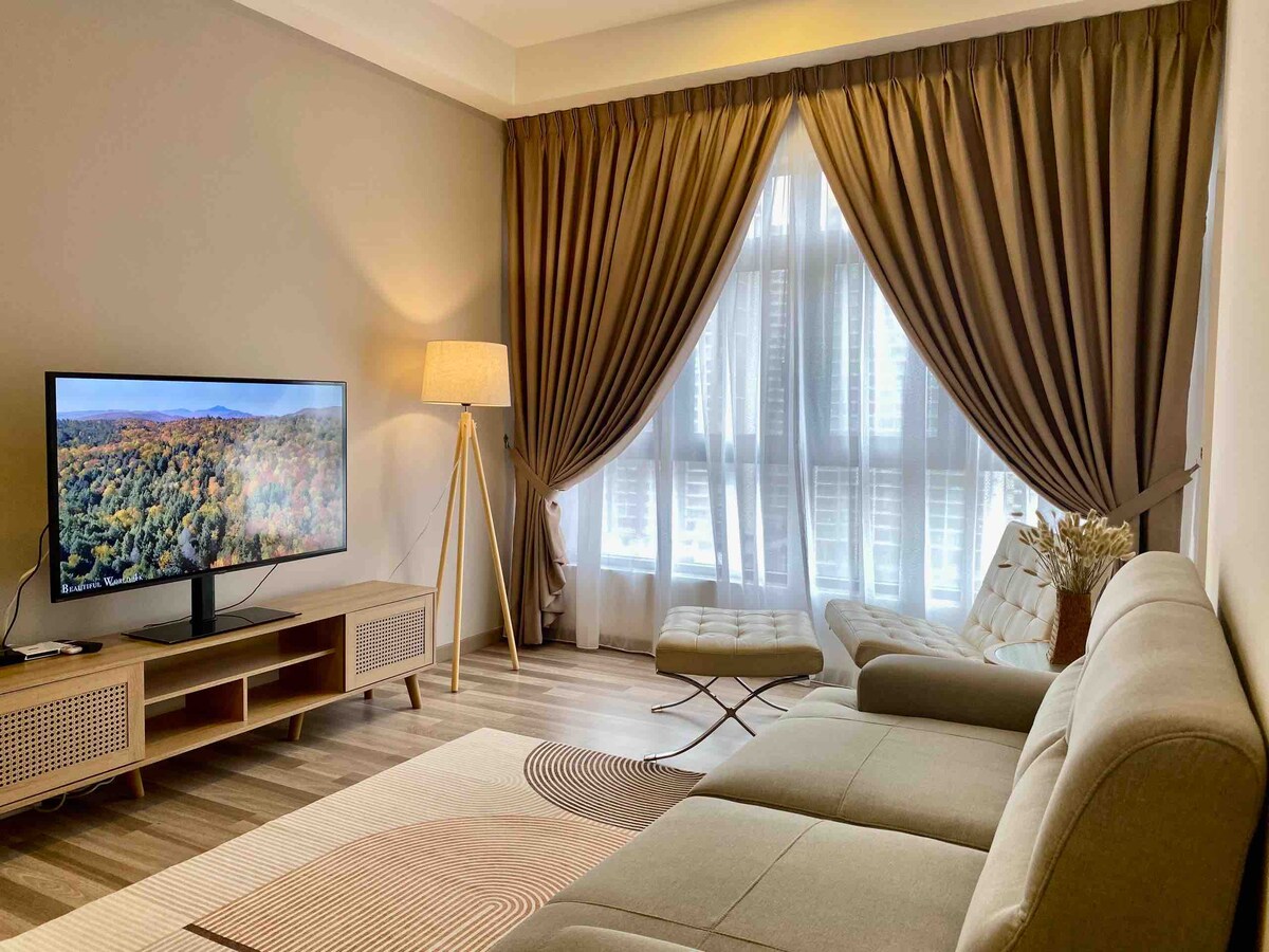 A modern living area features a comfortable sofa and a unique accent chair. A flat-screen TV is positioned on a clean-lined console. Natural light filters through large windows, adorned with elegant drapes, creating a warm ambiance across the wooden floor.