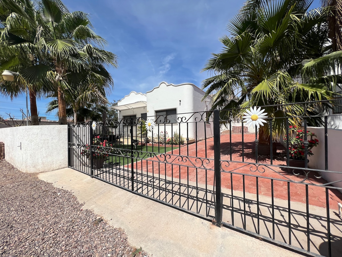 A gated entrance leads into the property, framed by vibrant green palm trees and flowering plants. The pathway is paved with red tiles, creating a welcoming transition to the house, which is visible in the background against a clear blue sky.