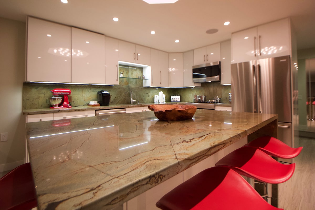 An open-concept kitchen features smooth, polished countertops with a rich stone pattern, complemented by sleek, modern cabinetry. Red bar stools are positioned at a spacious kitchen island, offering an inviting area for informal dining or gatherings.