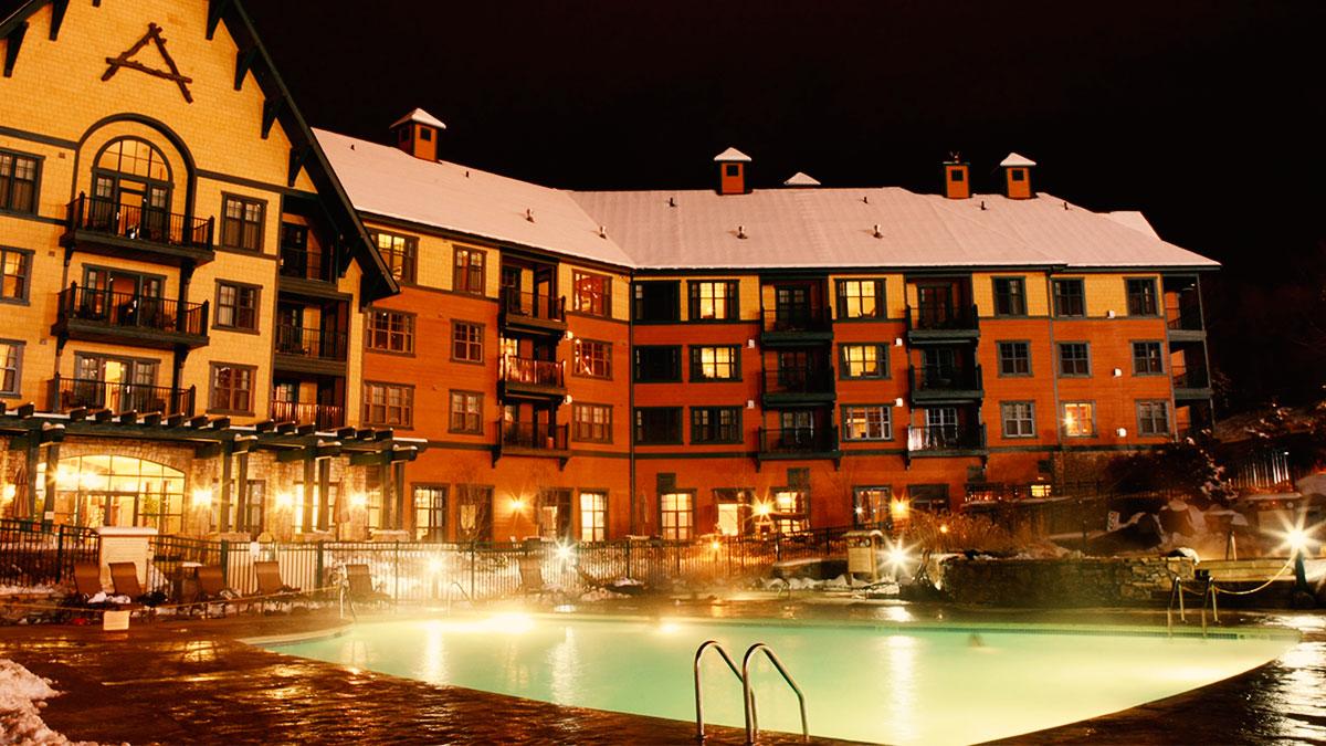 The exterior of The Appalachian Hotel is illuminated at night, showcasing the building's charming architecture. A spacious outdoor heated pool is visible in the foreground, surrounded by lounge chairs and snow-covered landscaping, contributing to the inviting ambiance.
