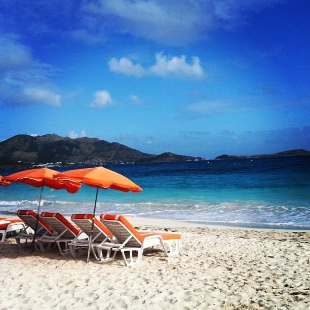 Several orange beach loungers are positioned under vibrant orange umbrellas, facing a calm sea. Gentle waves lap at the shore, where soft white sand is visible. Lush green hills outline the horizon, and a clear blue sky with scattered clouds adds to the serene coastal environment.
