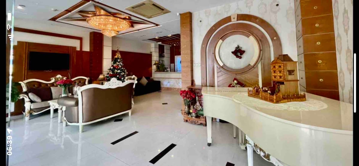 The lobby features a grand piano positioned beside elegantly arranged seating with a festive Christmas tree in the corner. Decorative accents enhance the space, and natural light illuminates the area through various window openings.
