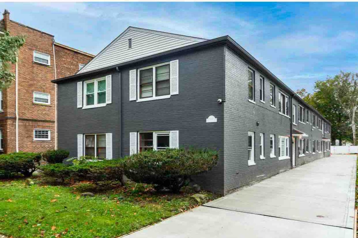 A two-story building is depicted, featuring a dark exterior with white trim. Windows are arranged symmetrically, allowing natural light to brighten the interiors. Lush hedges line the front, complemented by a paved walkway leading to the entrance, creating a welcoming approach.