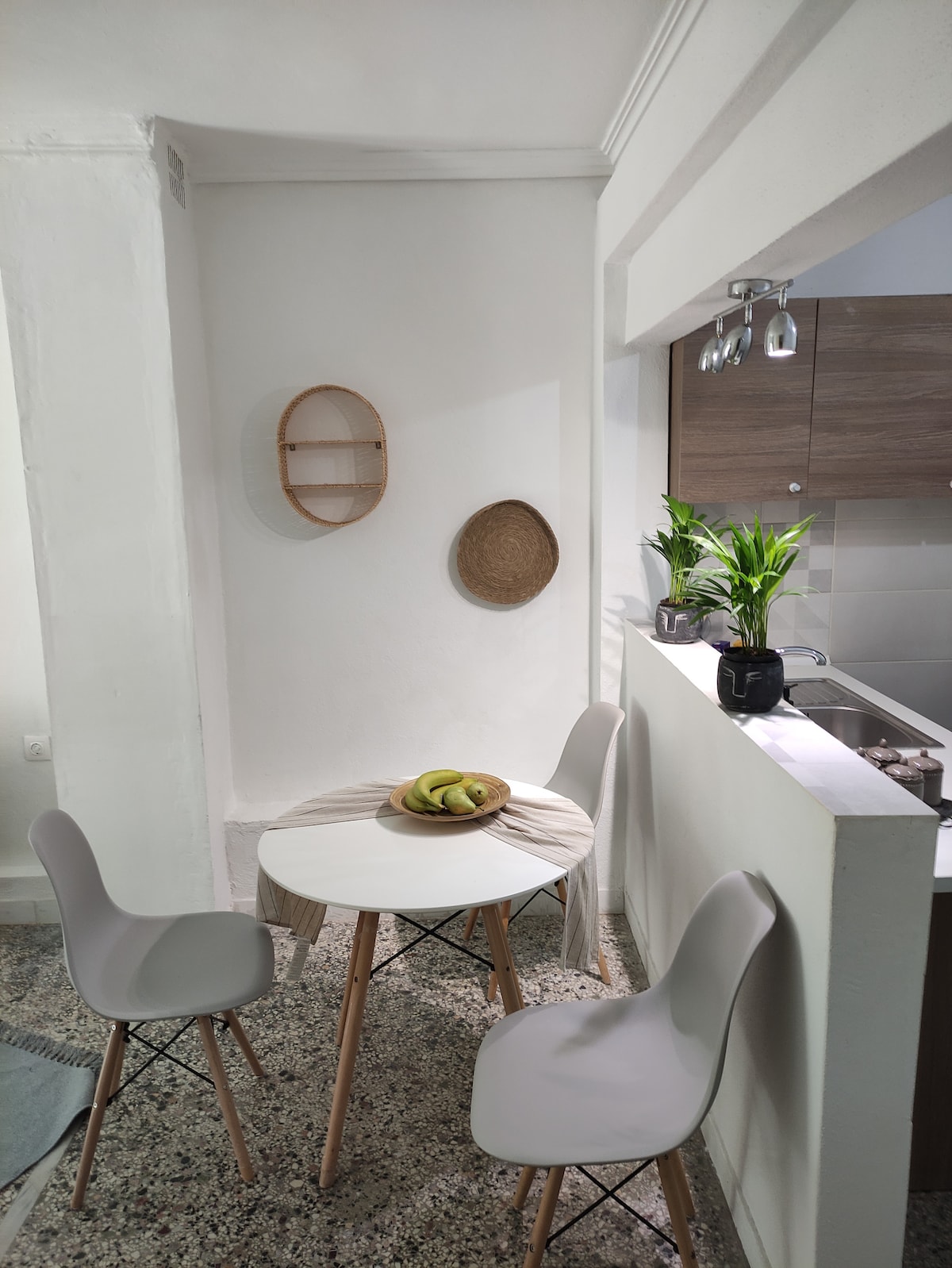 A dining area features a round table set with a bowl of bananas. Light-colored chairs surround the table, and a small shelf is mounted on the white wall. A potted plant adds a touch of greenery, and the kitchen area is visible in the background.