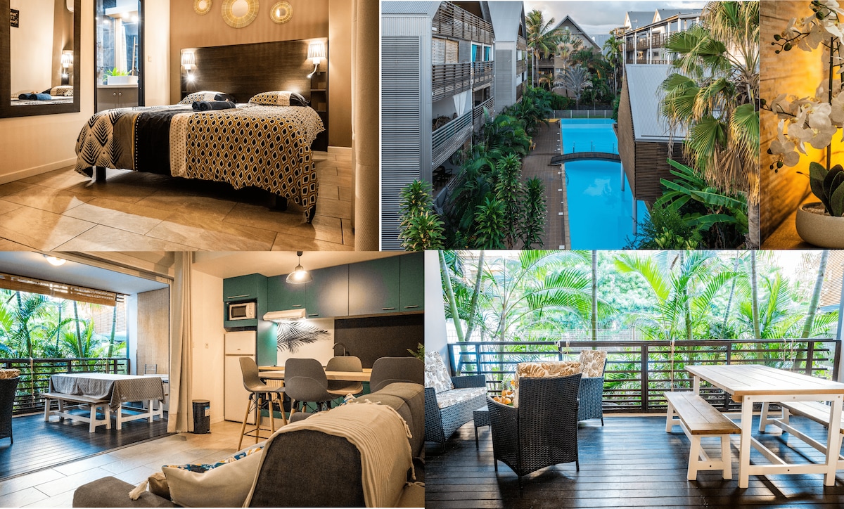 A spacious bedroom features a large bed with patterned bedding, soft lighting from wall fixtures, and a mirror on the wall. A living area includes a cozy couch and dining space, separated by a large sliding door leading to the terrace overlooking the pool and tropical greenery.