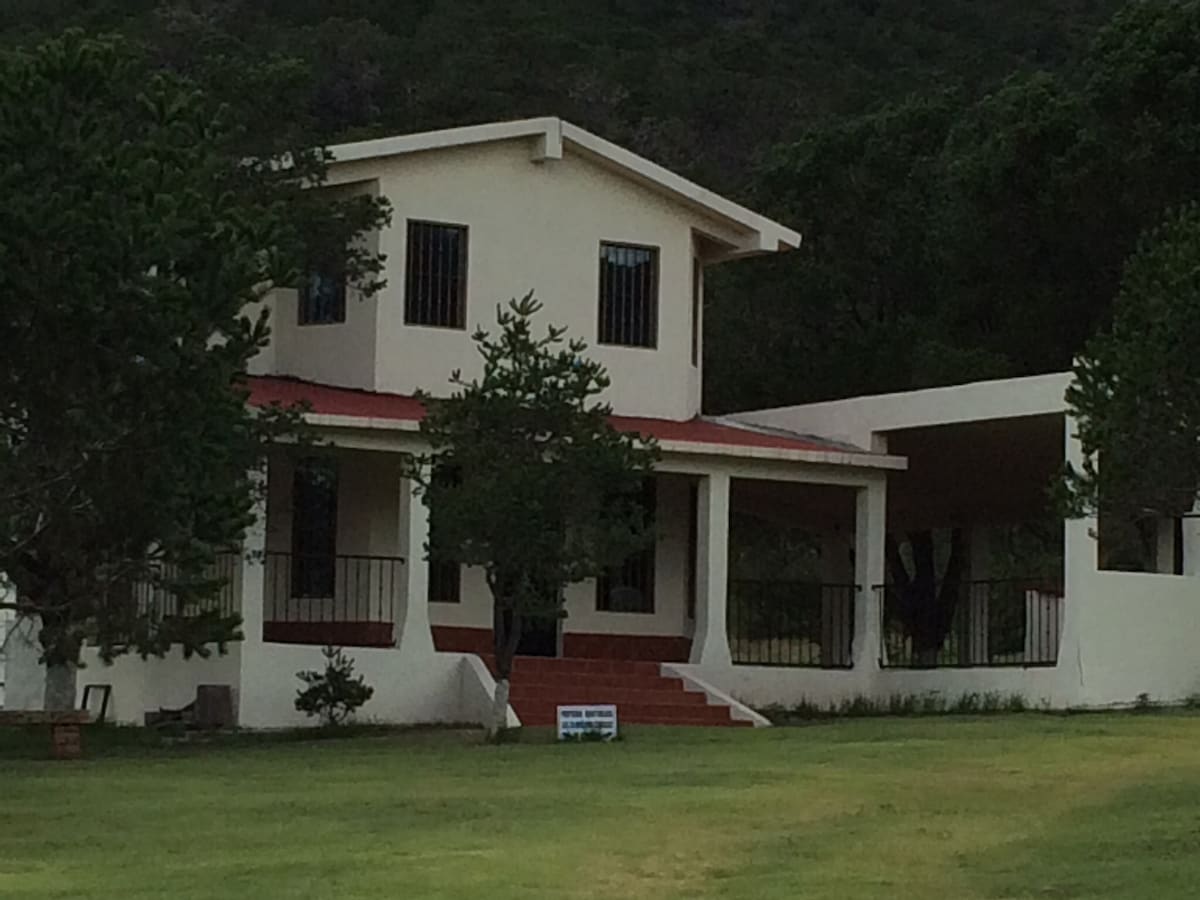 A spacious two-story house is set among a green landscape, featuring a red-tiled roof and tall windows. A staircase leads to the entryway, which is surrounded by greenery. An adjacent covered area offers additional outdoor space, providing a view of the surrounding hills.