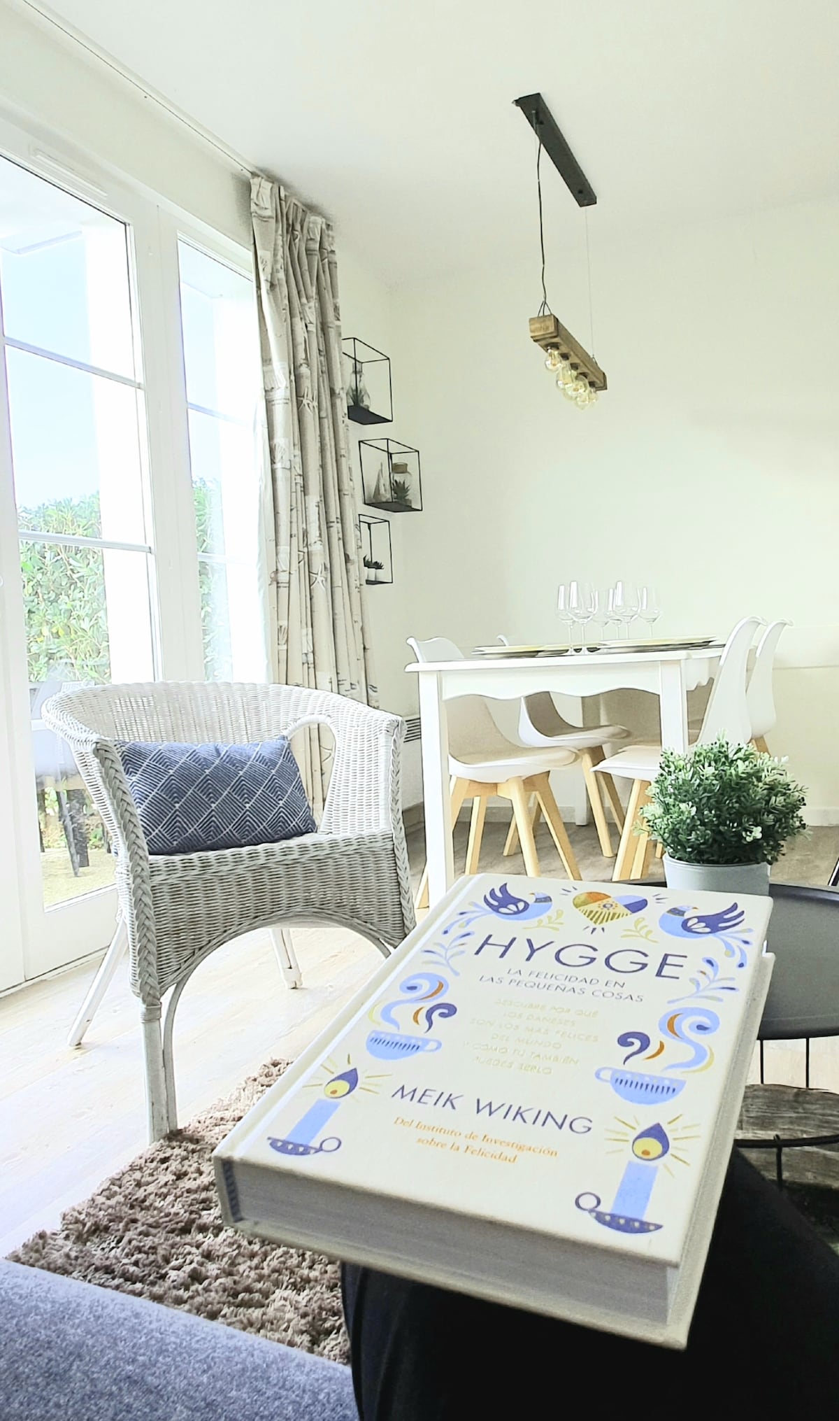 A bright and airy living space is showcased, featuring a cozy chair with a textured throw. A dining table with wooden chairs is seen alongside glass doors leading to an outdoor area. A decorative book lies on a textured surface, adding to the serene ambiance.