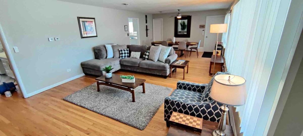 A spacious living area features a large gray sectional sofa and a patterned armchair. A wooden coffee table sits atop a soft area rug, while natural light filters through large windows. The open layout connects to a dining area, enhancing the communal atmosphere.