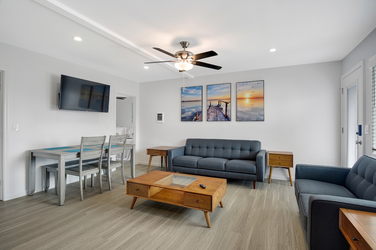 The living area showcases a modern design with hardwood flooring and a ceiling fan. A gray sofa and matching armchair are paired with a wooden coffee table. An entertainment unit holds a flat-screen TV, while a dining table is positioned nearby. Sunset-themed artwork adorns the wall.