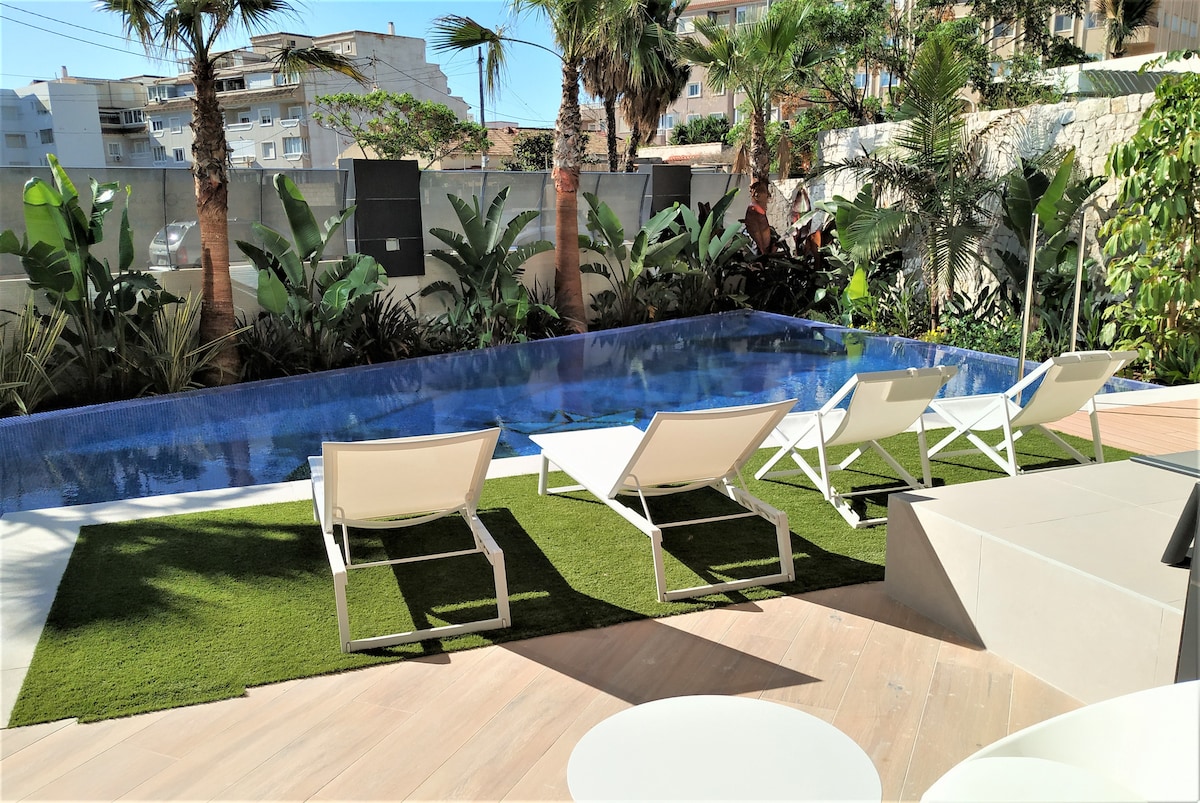 A serene outdoor area features a tranquil swimming pool surrounded by lush greenery. Several white lounge chairs are positioned on a grassy area, offering a comfortable spot to relax. The space is designed to invite moments of calm, with palm trees framing the view.