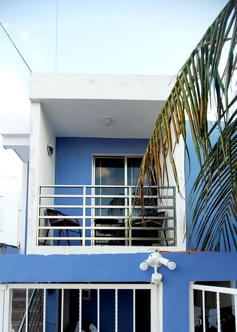 The exterior shows a two-story building with a blue façade. A balcony is visible on the upper level, featuring two chairs and a table, framed by a palm leaf. The entrance below includes a gated area with security lighting.