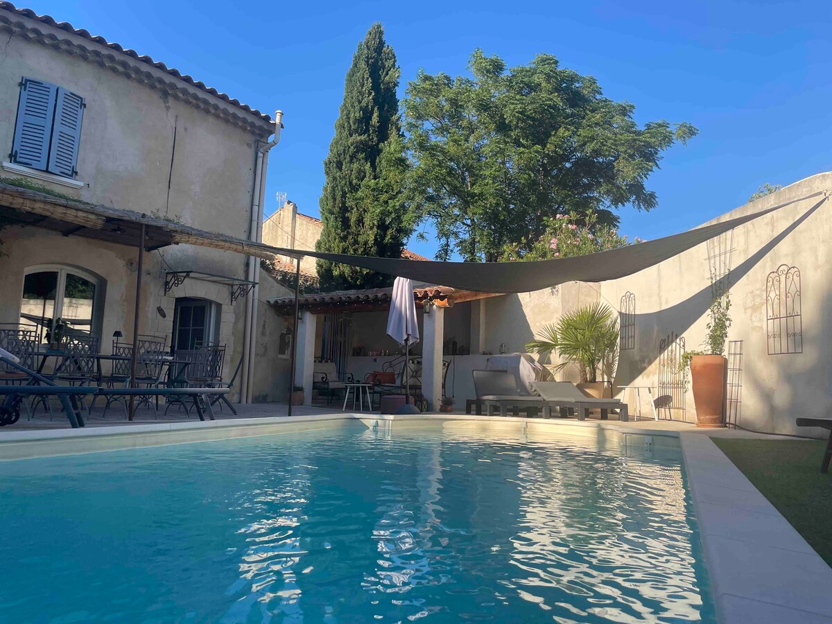 A view of the tranquil swimming pool is presented, surrounded by shaded seating areas and lush greenery. Sunlight casts reflections on the surface of the water. A canopy offers protection from the sun, while nearby plants add a touch of nature to the outdoor space.