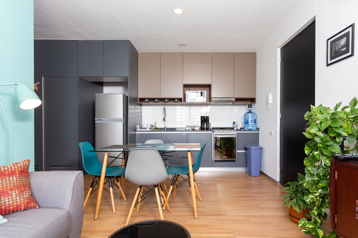 A modern kitchen and dining area is visible, featuring sleek cabinetry and essential appliances. A circular dining table with four chairs is set in the foreground, while a gray sofa adds comfort to the space. A water dispenser and potted plant bring a touch of practicality.