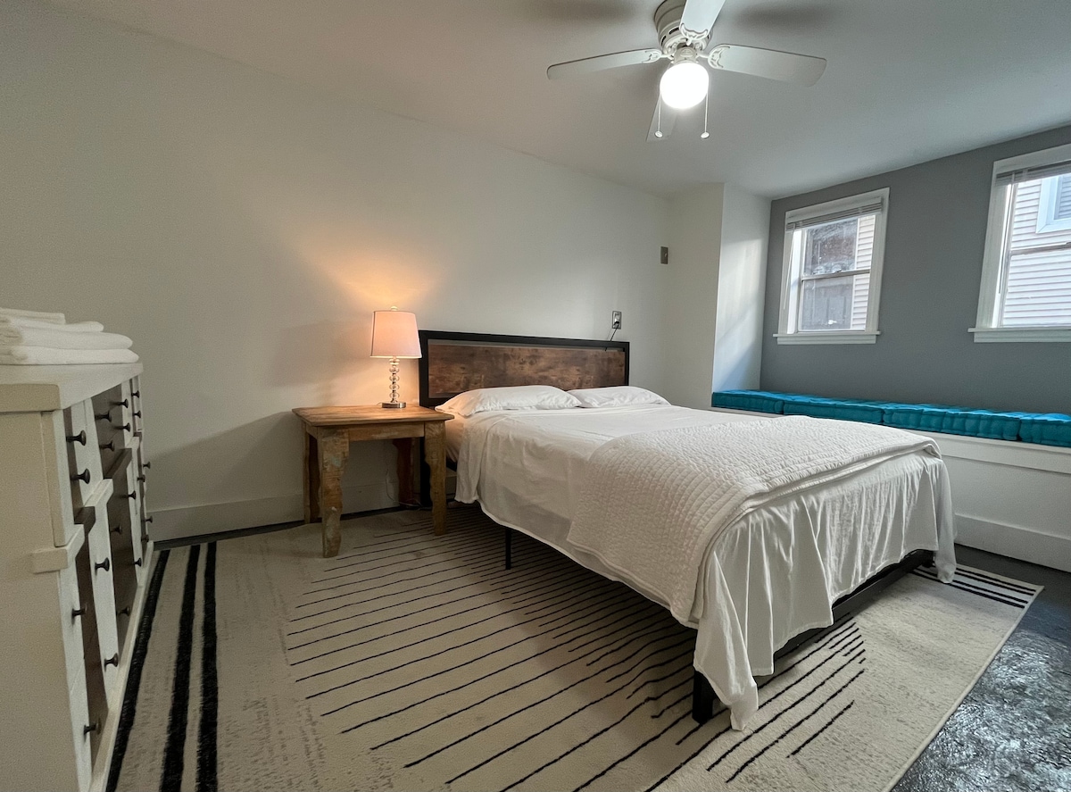 A well-lit sleeping area features a queen bed with crisp white bedding, complemented by a minimalist nightstand with a lamp. A futon sits nearby, and natural light filters through two windows, illuminating a patterned rug that anchors the space.