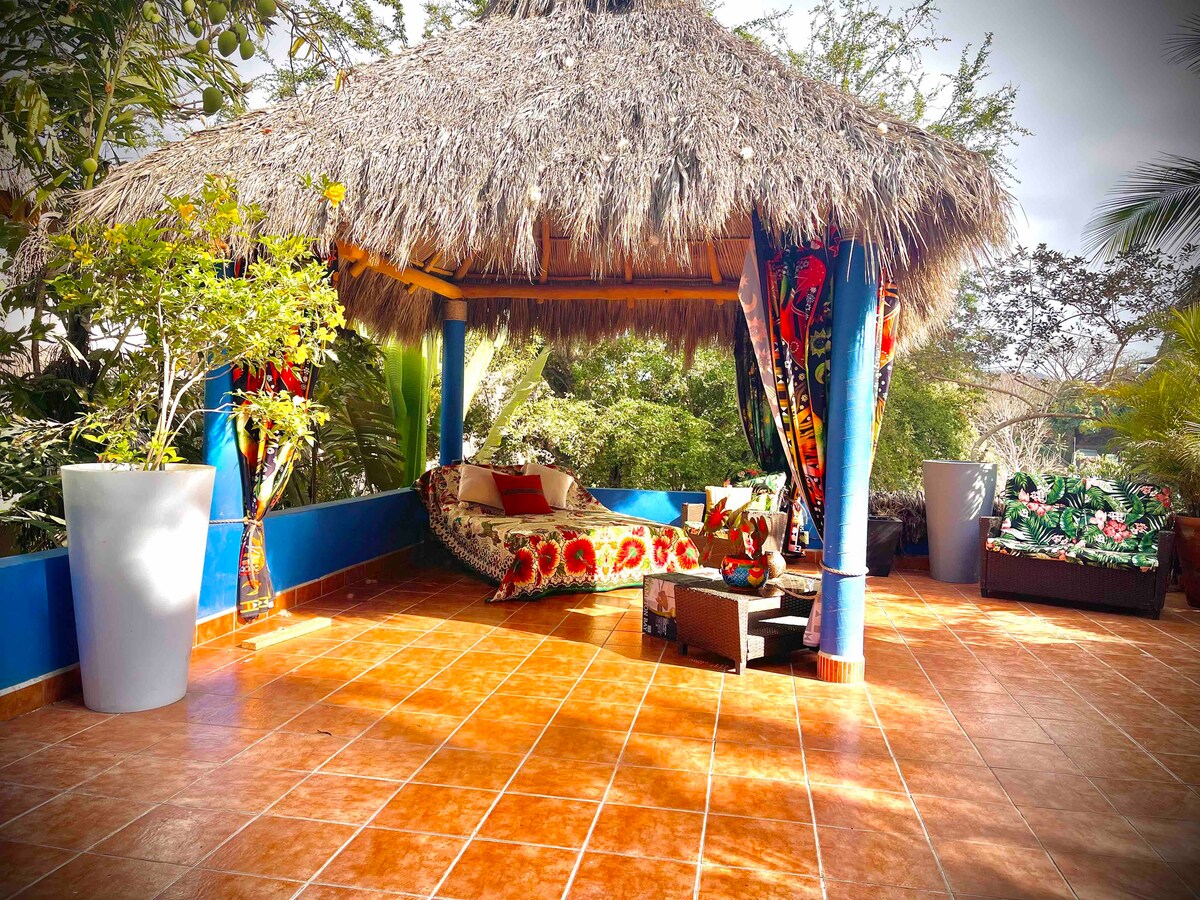 A spacious outdoor area is covered by a thatched palapa, featuring colorful fabric drapes. The tiled floor is complemented by decorative plants and seating, inviting relaxation in the lush surroundings. Natural light filters through the trees, enhancing the serene setting.