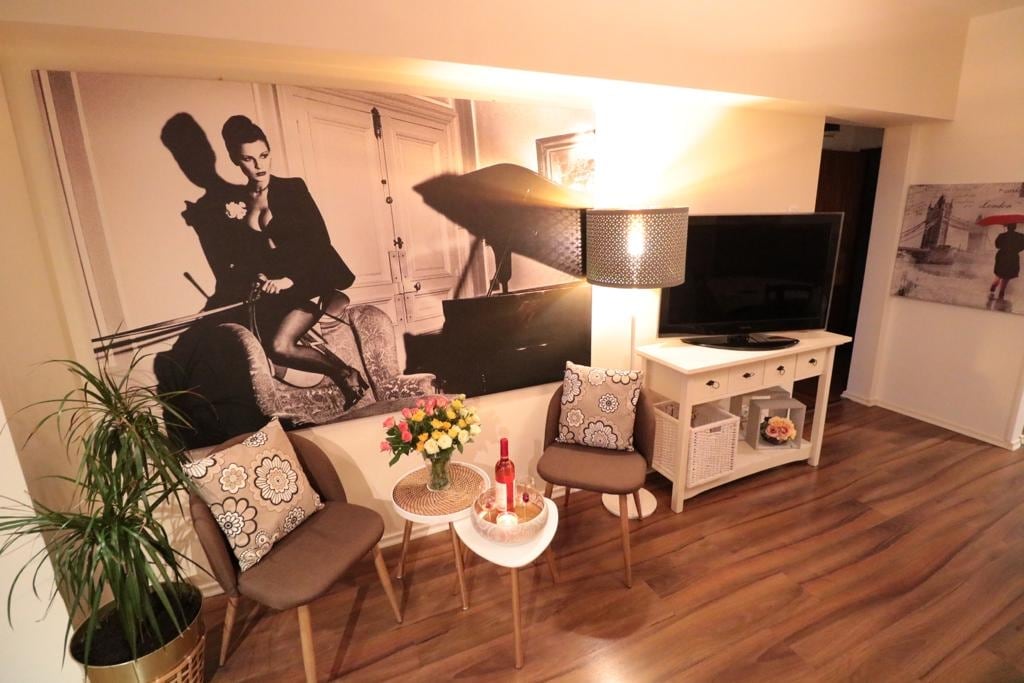 A stylish sitting area features two elegant chairs with patterned cushions, positioned around a small table. A floral arrangement sits atop the table. A large television is mounted on the wall, accompanied by a striking black and white mural, enhancing the space.