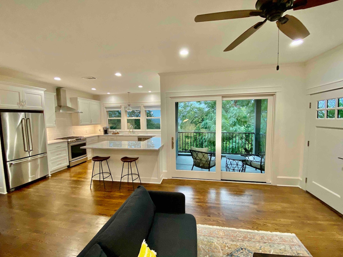 An open-concept living area features a modern kitchen with stainless steel appliances and a large island. Two bar stools are positioned at the island, and a comfortable black sofa faces sliding glass doors that lead to a balcony with greenery visible outside.