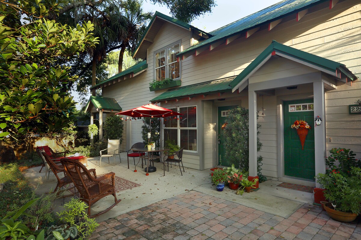 The exterior features a charming carriage house with a welcoming porch area surrounded by lush greenery. A small patio with tables and chairs is shaded by colorful umbrellas. Paved pathways and decorative plants enhance the inviting atmosphere of the entrance.